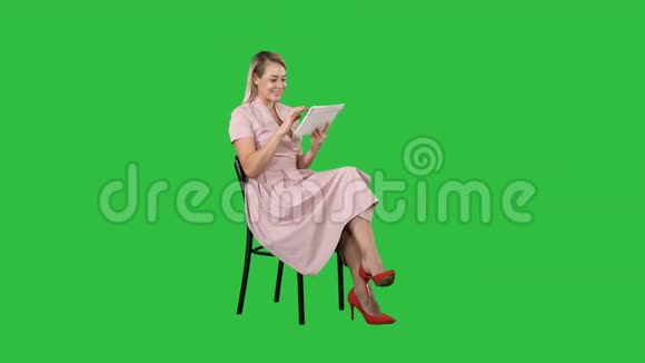 有趣的漂亮年轻女人穿着粉红色的连衣裙坐在椅子上用平板电脑在绿色屏幕上Chroma键视频的预览图