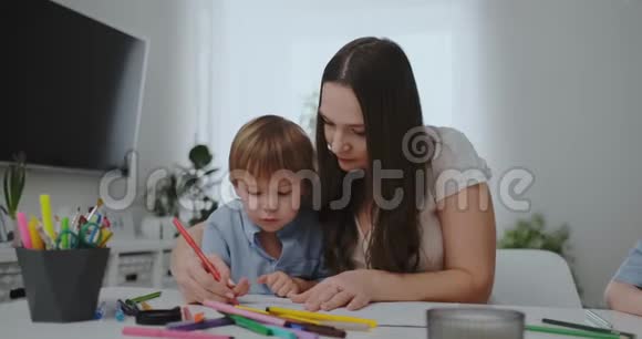 一家人有一个孩子一个男孩和一个年轻的母亲坐在桌子旁用彩色铅笔在纸上画画发展视频的预览图