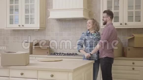 年轻的幸福家庭亲吻站在厨房在他们的新公寓穿格子衬衫戴眼镜的胡子老公视频的预览图