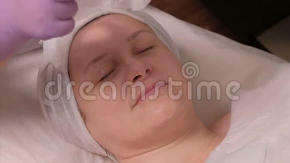 一位戴着紫丁香手套的专业美容师在一位中年妇女的脸上戴上面具皮肤再生的过程视频的预览图