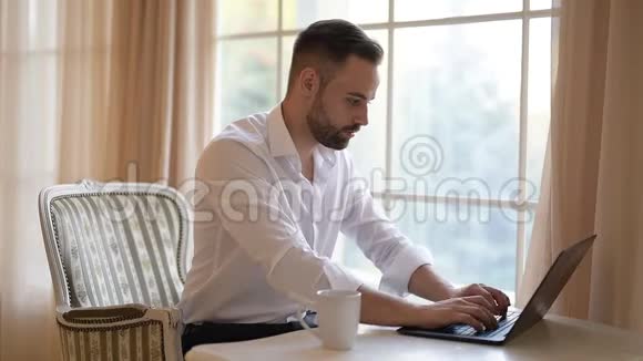 大胡子的商人正在喝咖啡在笔记本电脑的键盘上打字视频的预览图