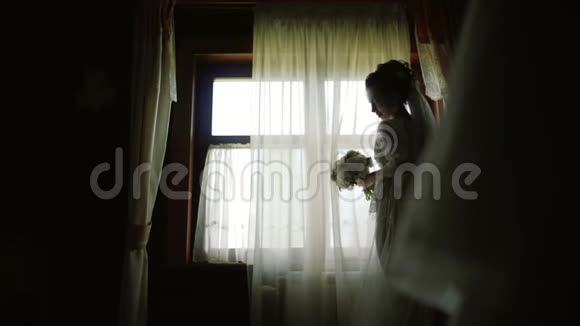 新娘穿着白色丝绸礼服在木房子的全景窗旁停留视频的预览图