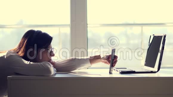 懒惰的年轻女性办公室工作人员戴着耳机上班时间玩手机游戏无聊视频的预览图