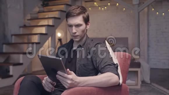 穿着黑色衬衫和牛仔裤的年轻帅哥坐在舒适的大椅子上看着数码平板电脑视频的预览图