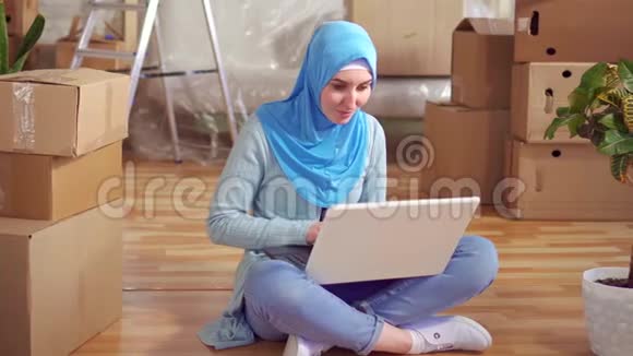 戴头巾的年轻穆斯林妇女使用笔记本电脑坐在盒子旁边的地板上视频的预览图