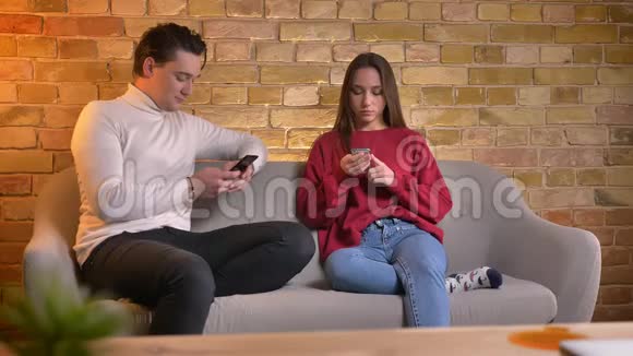 年轻的白种人夫妇坐在沙发上在家里的气氛中在智能手机上打字视频的预览图