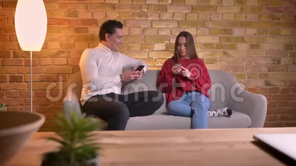 年轻的高加索夫妇坐在沙发上拿着智能手机在家里的气氛中互相交谈视频的预览图