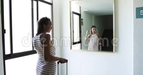 一个年轻的女孩穿着一件连衣裙车轮上有一个手提箱在酒店大厅里看着镜子视频的预览图