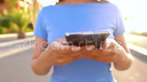 日落时分女人在棕榈街散步时使用智能手机视频的预览图
