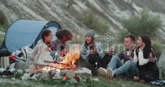 在露营的不同种族微笑有魅力的朋友坐在篝火旁喝一些热饮视频的预览图