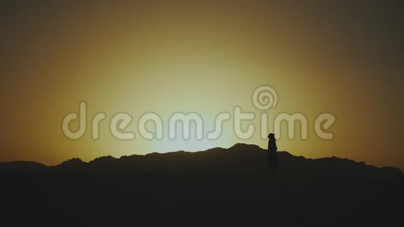 美丽的年轻女子在沙漠中日落时散步的剪影穿过沙漠沙丘风吹缓慢视频的预览图
