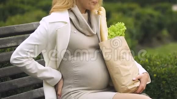 年轻孕妇带着购物袋在长凳上休息背部疼痛视频的预览图