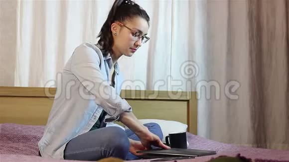 一个漂亮的年轻女孩正在打开她的笔记本电脑在坐在床上的键盘上打字视频的预览图