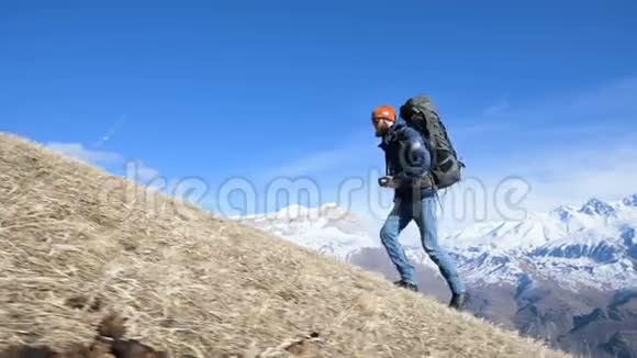 一位留胡子的年轻男性摄影师戴着一顶帽子和太阳镜背着一个大背包脖子上戴着一台照相机爬上了山顶视频的预览图
