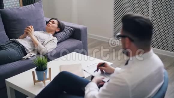 心烦意乱的女孩和躺在办公室沙发上的心理学家讨论情绪视频的预览图