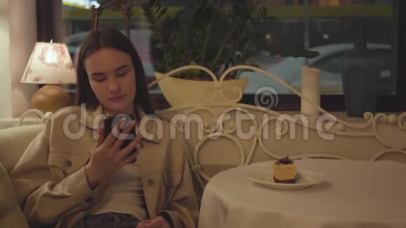 漂亮的女孩坐在餐馆的桌子上用手机发短信这块美味的蛋糕在桌子上视频的预览图