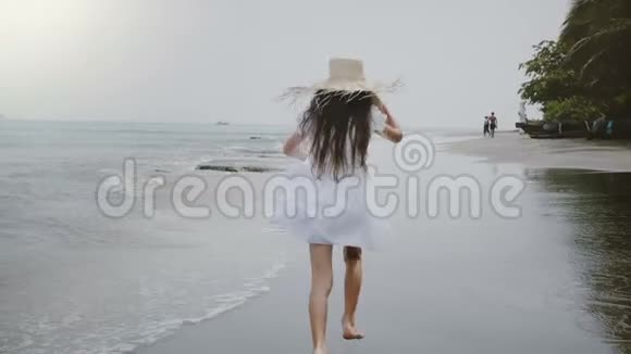 镜头跟随快乐美丽的小女孩长发和草帽沿着大气热带海滩奔跑视频的预览图