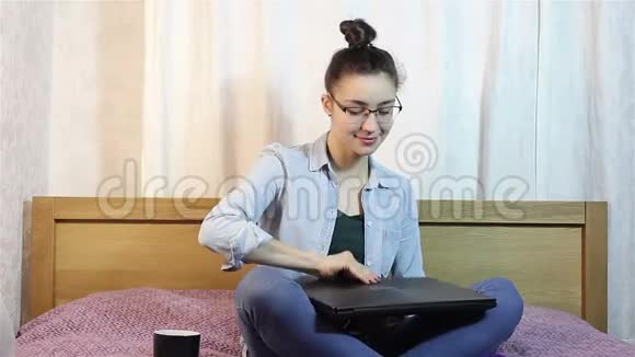 一个漂亮的年轻女孩正在打开她的笔记本电脑在坐在床上的键盘上打字视频的预览图