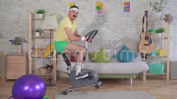 有趣的精力充沛的80多岁男人带着胡子在家里骑着运动自行车慢悠悠视频的预览图