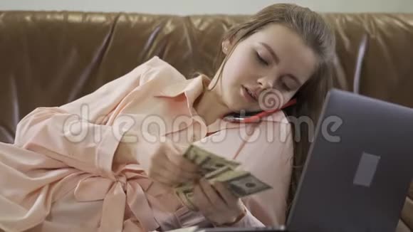 年轻漂亮的女孩坐在真皮沙发上用手机说话看着电脑数着钱的现金视频的预览图