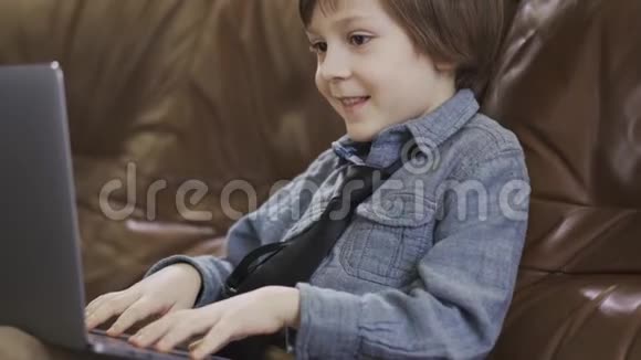 穿着牛仔裤夹克的小男孩坐在皮革沙发上玩着笔记本电脑开心地笑着现代的休闲视频的预览图