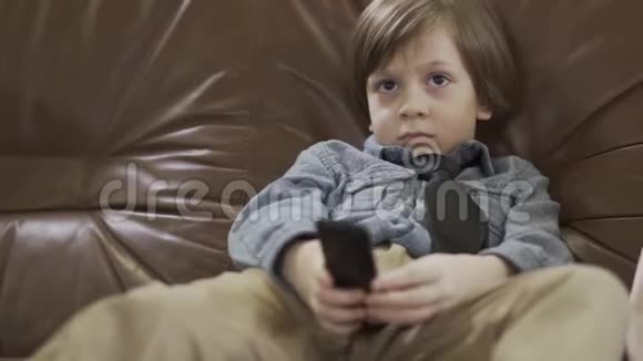 这个小可爱的男孩坐在皮革沙发上两腿分开用遥控器在电视上换频道可爱的孩子视频的预览图