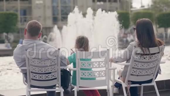 快乐的父亲母亲和女儿坐在城市公园的喷泉前视频的预览图