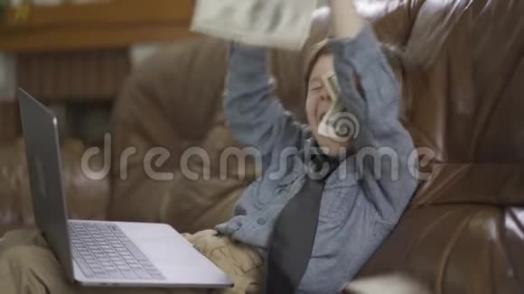 肖像小男孩坐在皮革沙发上腿上有笔记本电脑在空中扔钱金钱视频的预览图