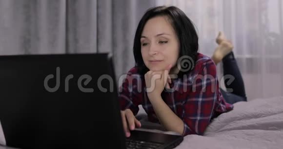 年轻的黑发女人躺在床上聊天在笔记本电脑上工作视频的预览图