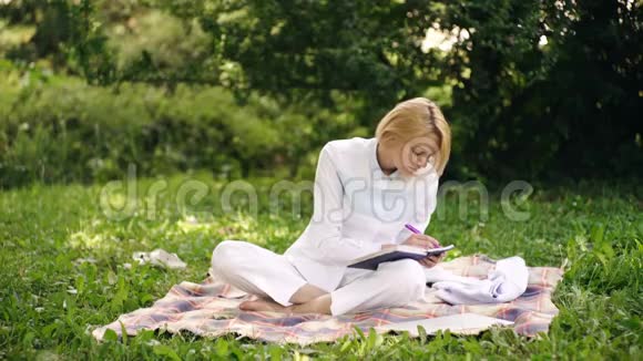快乐的年轻女人在公园里用笔记本电脑工作坐在公园里的女人拿着笔记本电脑和笔记本电脑坐在绿草上魅力视频的预览图
