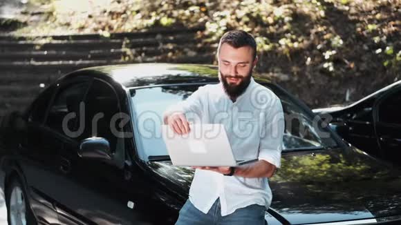 穿着衬衫的胡子男在汽车附近使用笔记本电脑视频的预览图