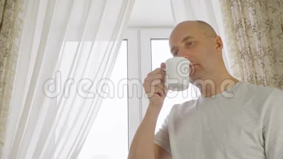 在窗户背景的舒适厨房里成人男子正在喝早咖啡一个喜欢喝热茶的人视频的预览图