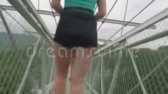 两个赤着双腿戴着虚拟头盔的白种人女孩正沿着绿色森林上的高吊桥行走关闭视频的预览图