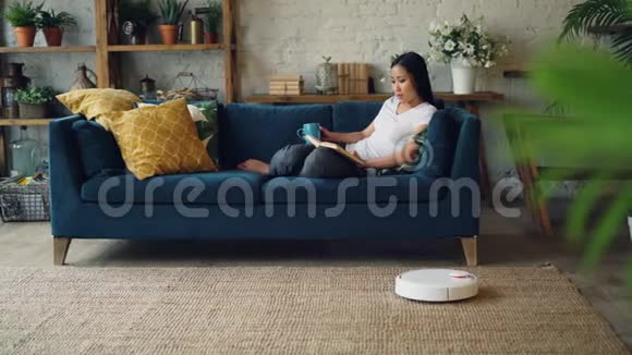 放松的亚洲女人正在看书在家里坐在沙发上翻书页机器人胡佛正在打扫地板视频的预览图