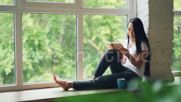 迷人的亚洲女学生正在阅读书籍微笑着坐在现代公寓的窗台上爱好青年视频的预览图