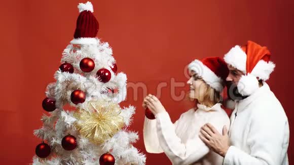 戴红帽子的长胡子男人拥抱一个装饰圣诞树的女人可爱的夫妇装饰圣诞树视频的预览图