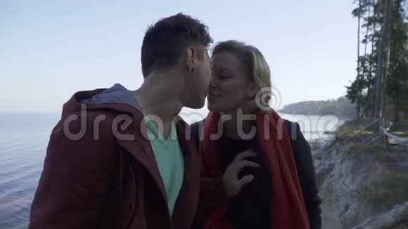 美丽的年轻多民族微笑的情侣在悬崖松树和湖泊的背景下相爱接吻视频的预览图