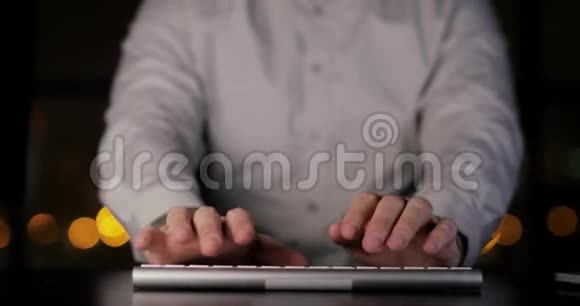 面目全非的男人正在黑暗中坐在工作场所的桌子旁的键盘上打字双手特写视频的预览图
