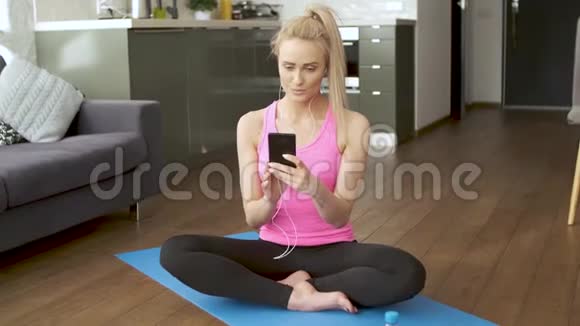 可爱的女人坐在瑜伽垫上浏览手机唱歌视频的预览图