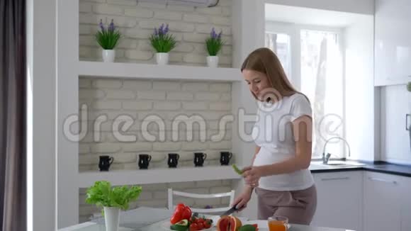 漂亮的怀孕女孩站在餐桌旁手里拿着刀子吃和切蔬菜视频的预览图