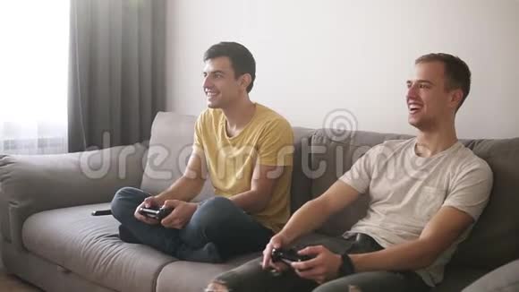 两个年轻人在家玩电子游戏拿着操纵杆坐在阁楼内部房间的灰色沙发上微笑视频的预览图