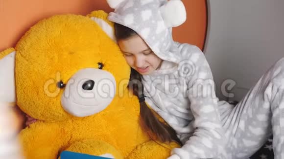十几岁的女孩穿着温暖的睡衣和一个朋友在手机上聊天而坐在床上和一只泰迪熊视频的预览图