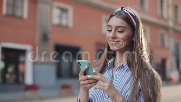 微笑的年轻女子穿着蓝色和白色条纹连衣裙用智能手机在老街上走来走去视频的预览图