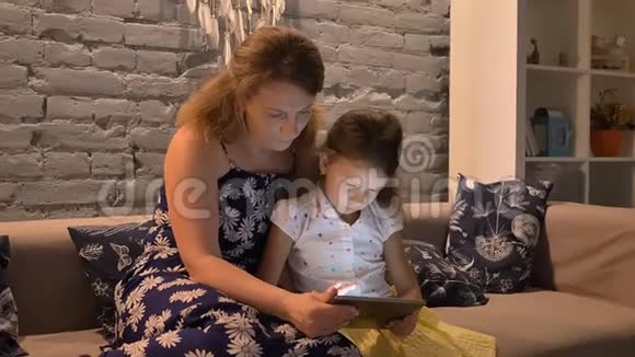 妈妈和小女儿坐在沙发上用平板电脑妈妈帮助孩子做小玩意家庭观念室内视频的预览图