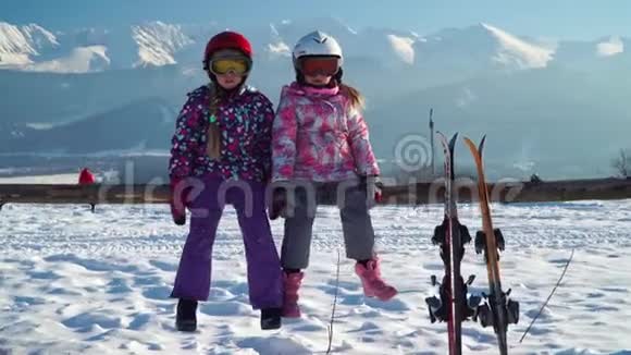 穿着外套戴着头盔的小女孩坐在木栅栏上滑雪板靠近群山视频的预览图