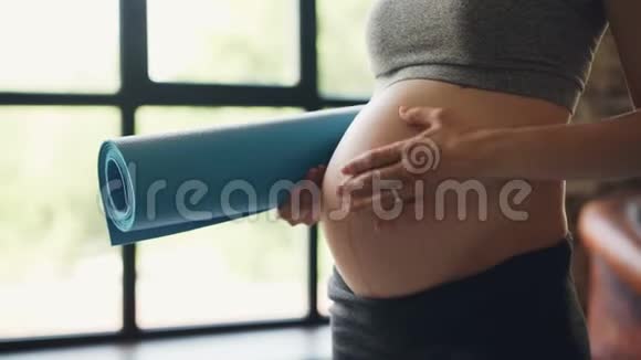 孕妇腹部特写镜头和妇女手抚摸肚子而另一只手拿着瑜伽垫健康生活方式视频的预览图