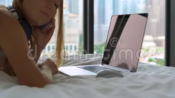 一名躺在床上的年轻女子在听到音乐时使用无线耳机在笔记本电脑上听音乐的特写镜头视频的预览图