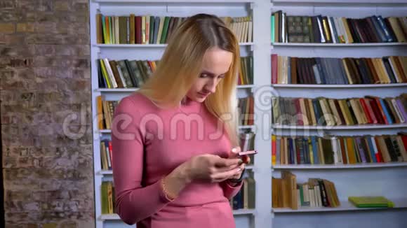金发白种人年轻女孩在书架背景上专心看智能手机的肖像视频的预览图