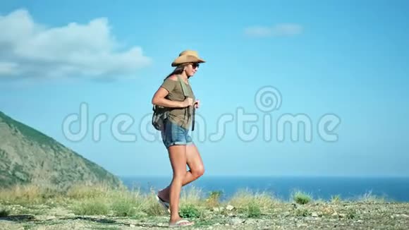 背包客徒步旅行的女人戴着太阳镜戴着帽子走在山边的海边风景视频的预览图