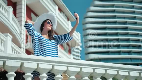 戴着帽子和太阳镜的魅力女人在酒店露台上用智能手机自拍视频的预览图